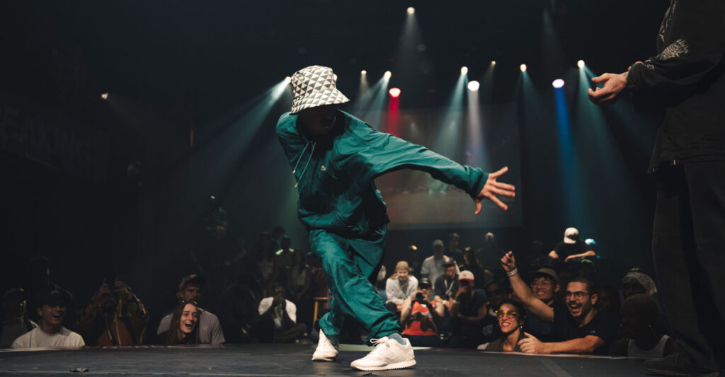 Un homme qui danse sur une scène, il a un autre homme en face de lui qui attend son tour de danser. Il a une foule de personne qui encourage et regarde la personne qui danse.