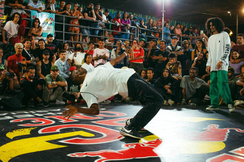 Il y a un homme qui danse dans un cercle entouré d'une foule de personne qui le regarde, il a un autre homme qui attend son tour pour pouvoir lui danser à son tour.